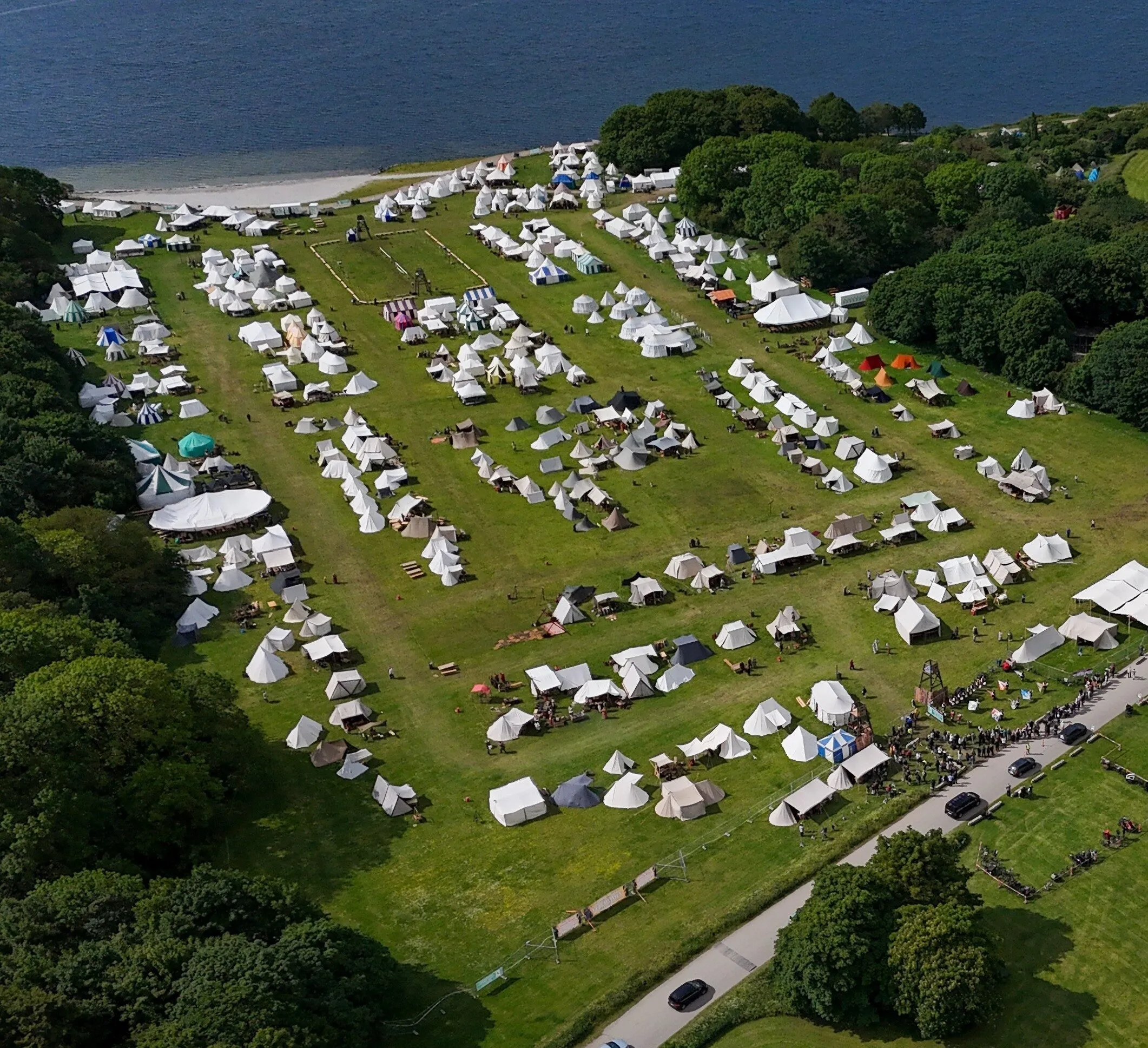 Dronefoto af markedspladsen i Valbyparken med rækker af hvide og stribede telte på græsplænen, turneringsbanen i midten, skov på begge sider og vandet i baggrunden